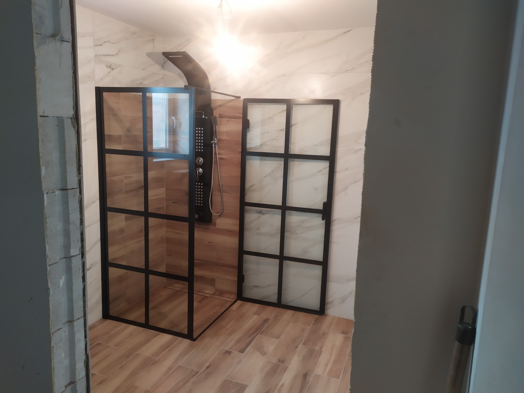 Kabina prysznicowa typu walk-in z czarnymi profilami, zamontowana w łazience z marmurowymi płytkami, widoczny surowy fragment ściany przed wykończeniem.