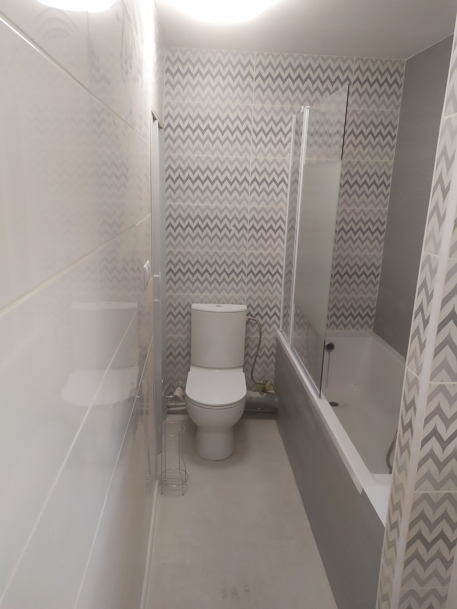 Wąska łazienka po remoncie, z toaletą, wanną z parawanem nawannowym i płytkami w zygzaki w odcieniach szarości i bieli.