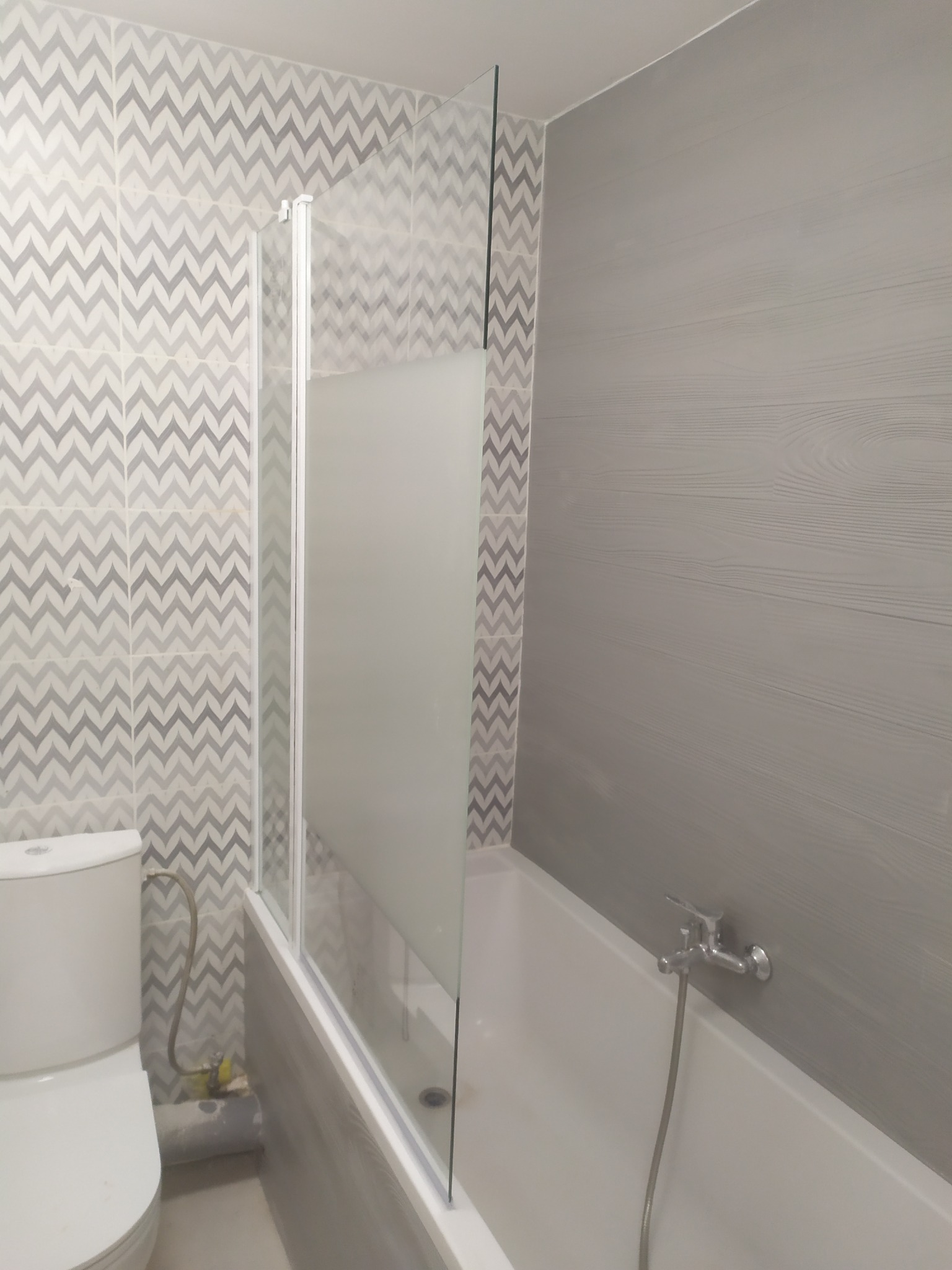 Szaro-biała łazienka po remoncie z wanną, toaletą i szklanym parawanem nawannowym, wzorzyste płytki chevron na ścianie i drewnopodobne panele ścienne, widoczne rury doprowadzające wodę do toalety.