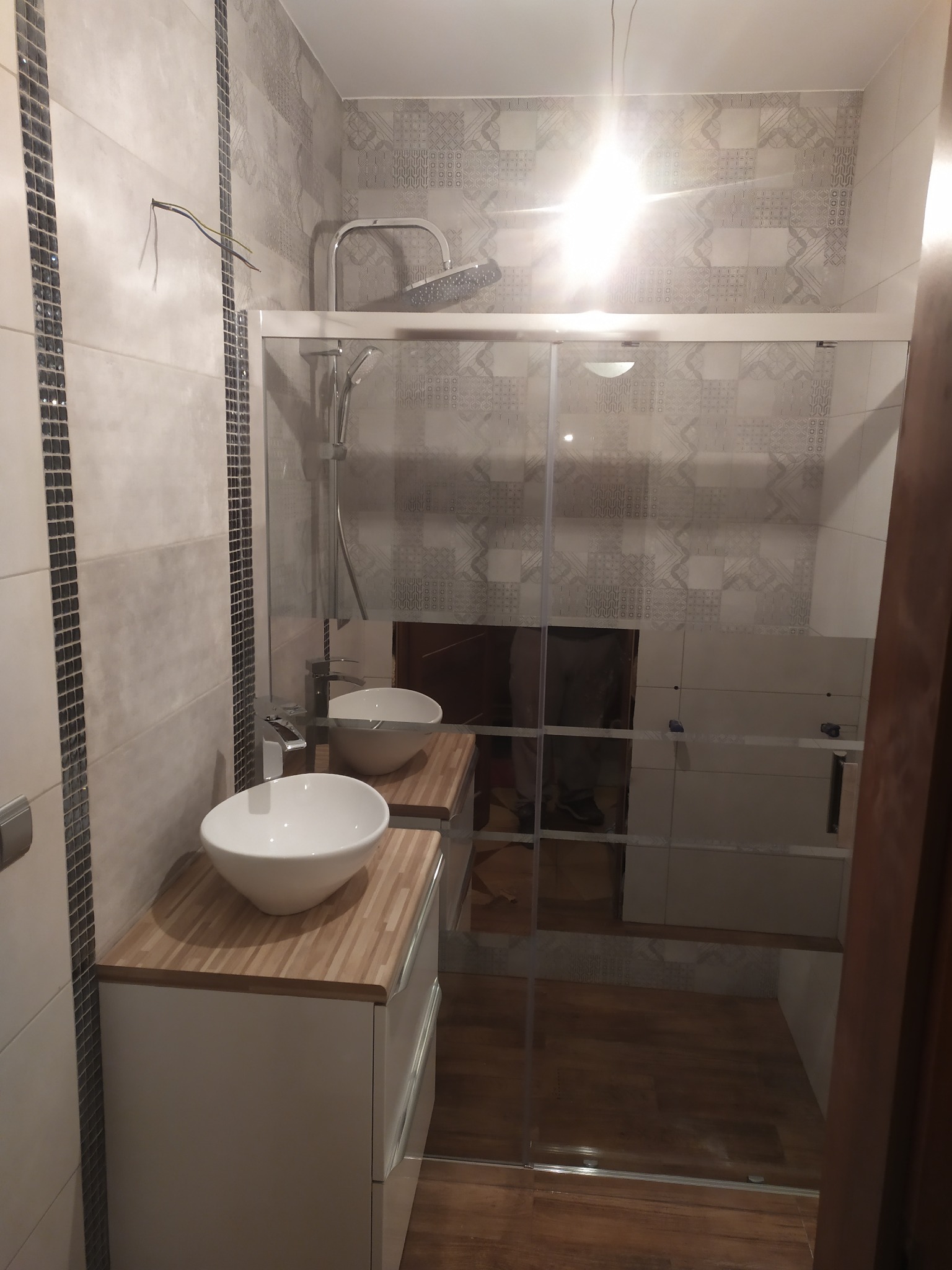 Nowoczesna łazienka w trakcie wykańczania: umywalki nablatowe na drewnianych blatach, przeszklona kabina prysznicowa, płytki w geometryczne wzory, wystające przewody elektryczne.