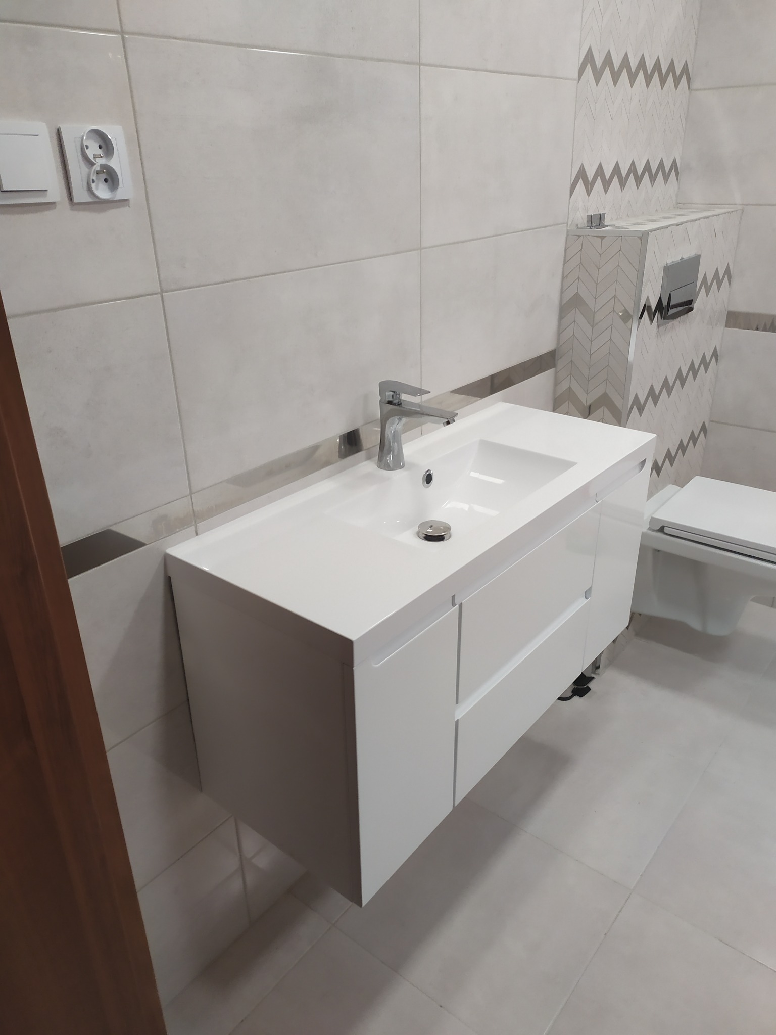 Nowoczesna, biała umywalka z szafką i chromowaną baterią w łazience z jasnymi płytkami i dekoracyjnym pasem z motywem zygzaków.