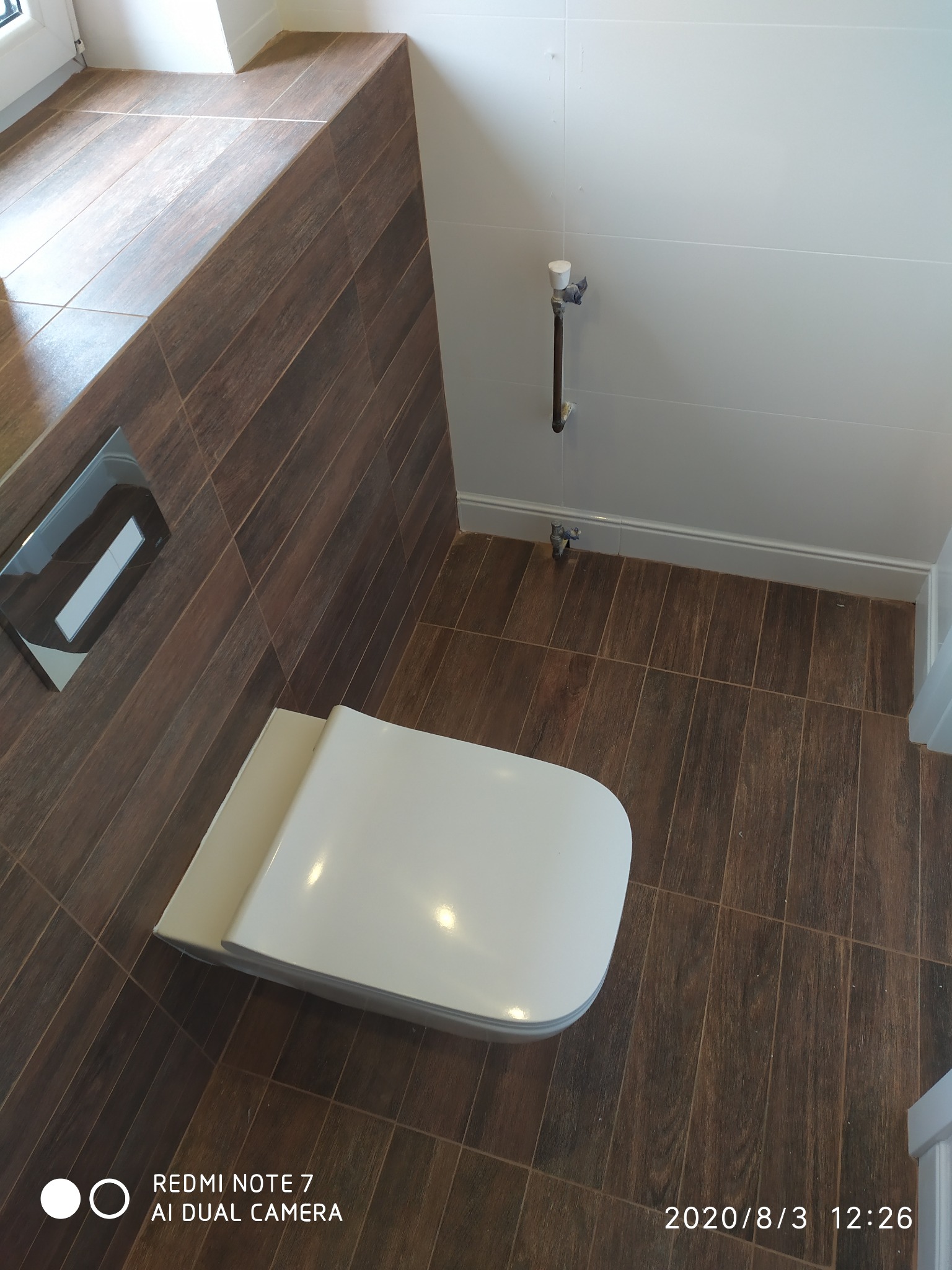 Nowoczesna toaleta podwieszana z brązowymi płytkami imitującymi drewno na podłodze i ścianie, widoczne zawory na ścianie z białymi płytkami.