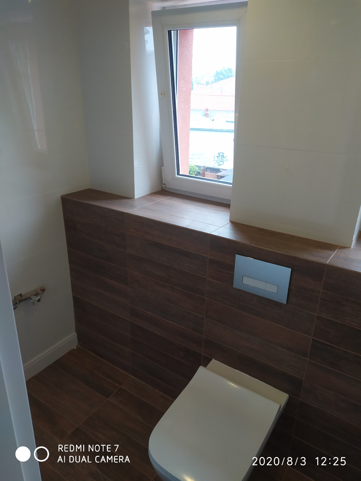 Nowoczesna toaleta podwieszana z brązowymi płytkami imitującymi drewno, białe płytki na ścianach i okno z widokiem na zewnątrz.