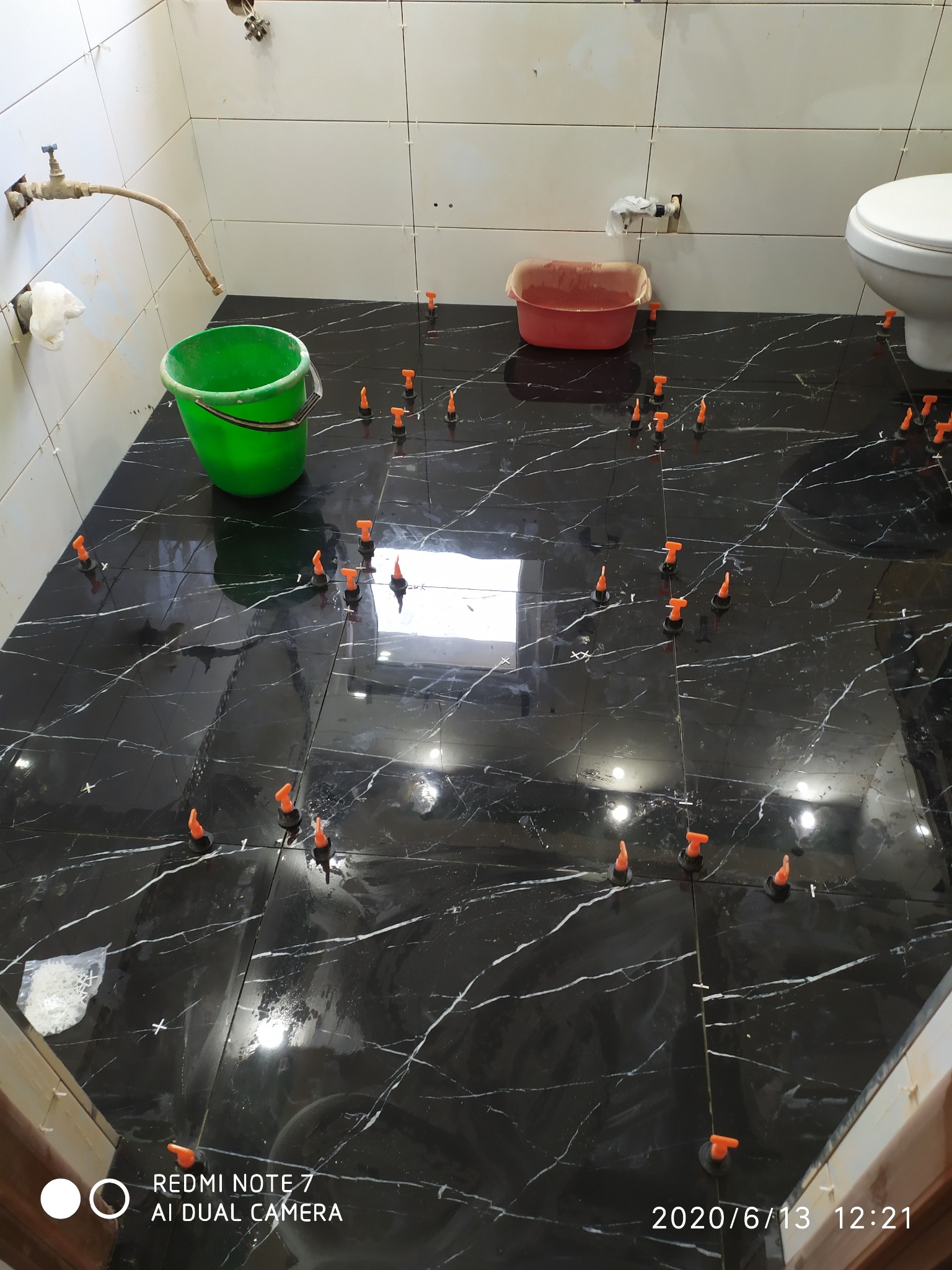 Układanie czarnych płytek podłogowych z białym żyłowaniem w łazience, z użyciem pomarańczowych klipsów poziomujących i zielonego wiadra.