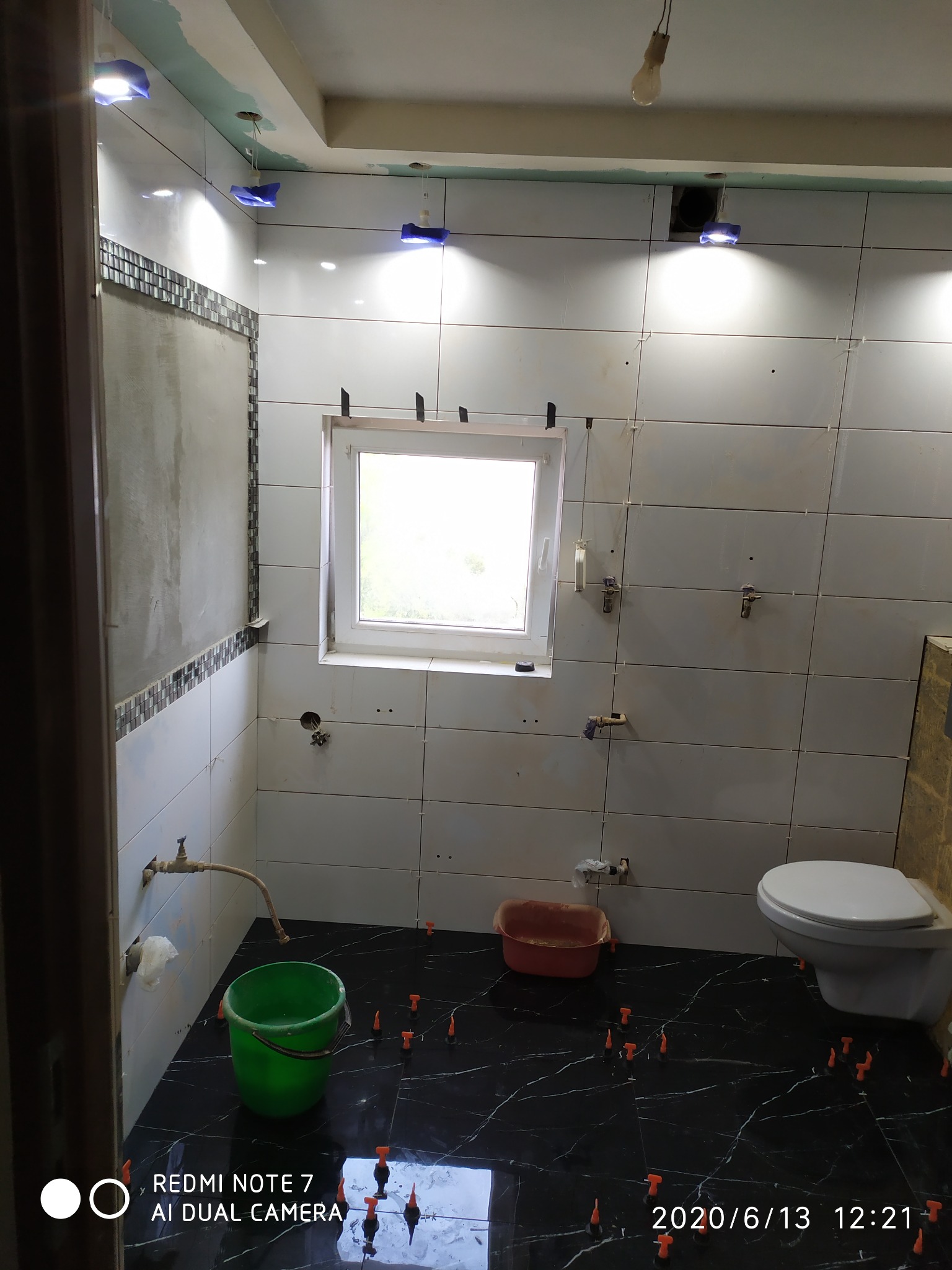 Łazienka w trakcie układania płytek: białe płytki ścienne z poziomymi szarymi paskami dekoracyjnymi, czarna marmurkowa podłoga, widoczne poziomice i plastikowe krzyżyki, surowe oświetlenie, okno...