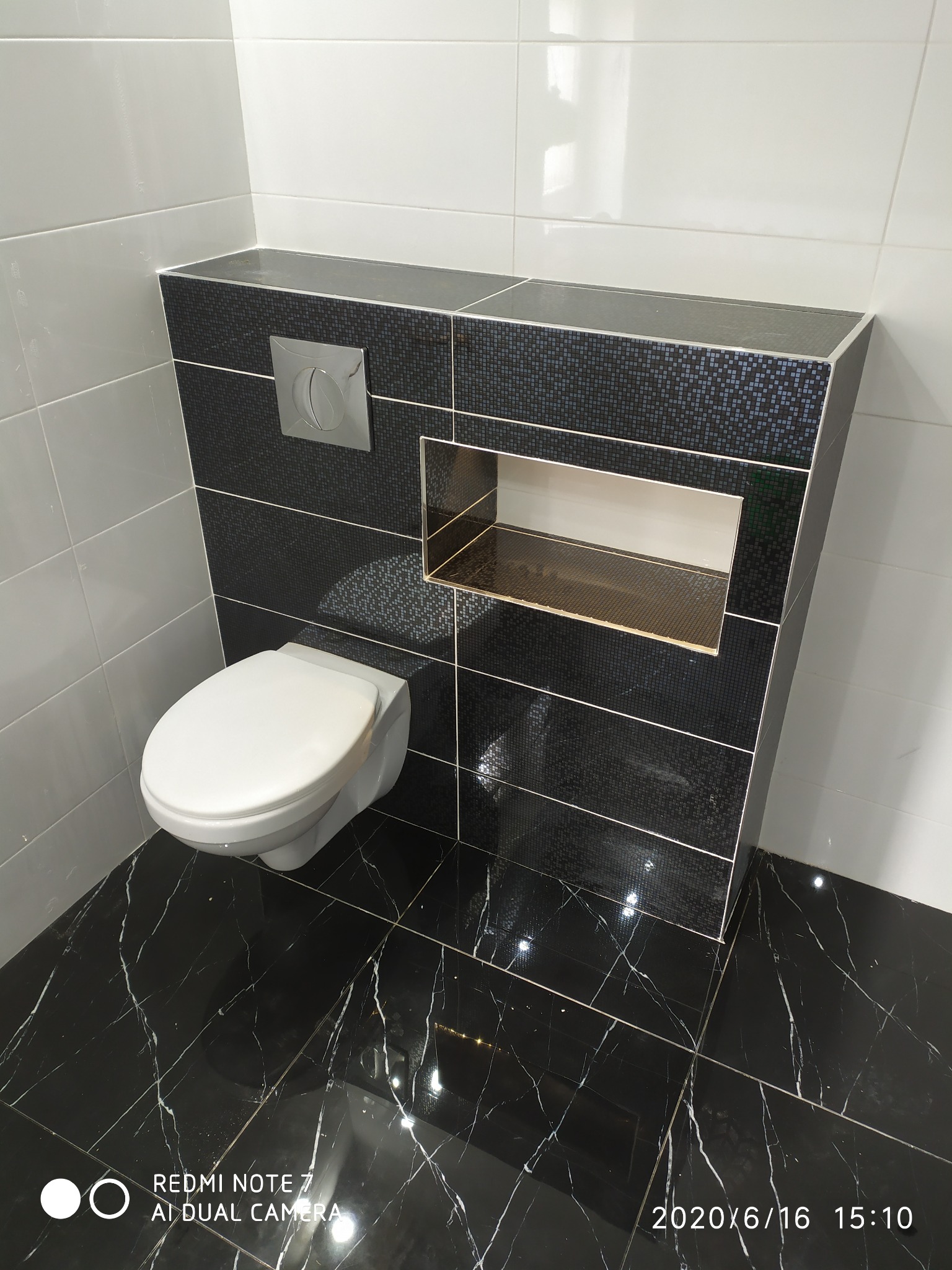 Nowoczesna toaleta podwieszana z czarną mozaiką i wnęką, czarna marmurowa podłoga z białymi żyłami, białe płytki na ścianie, chromowany przycisk spłukiwania.