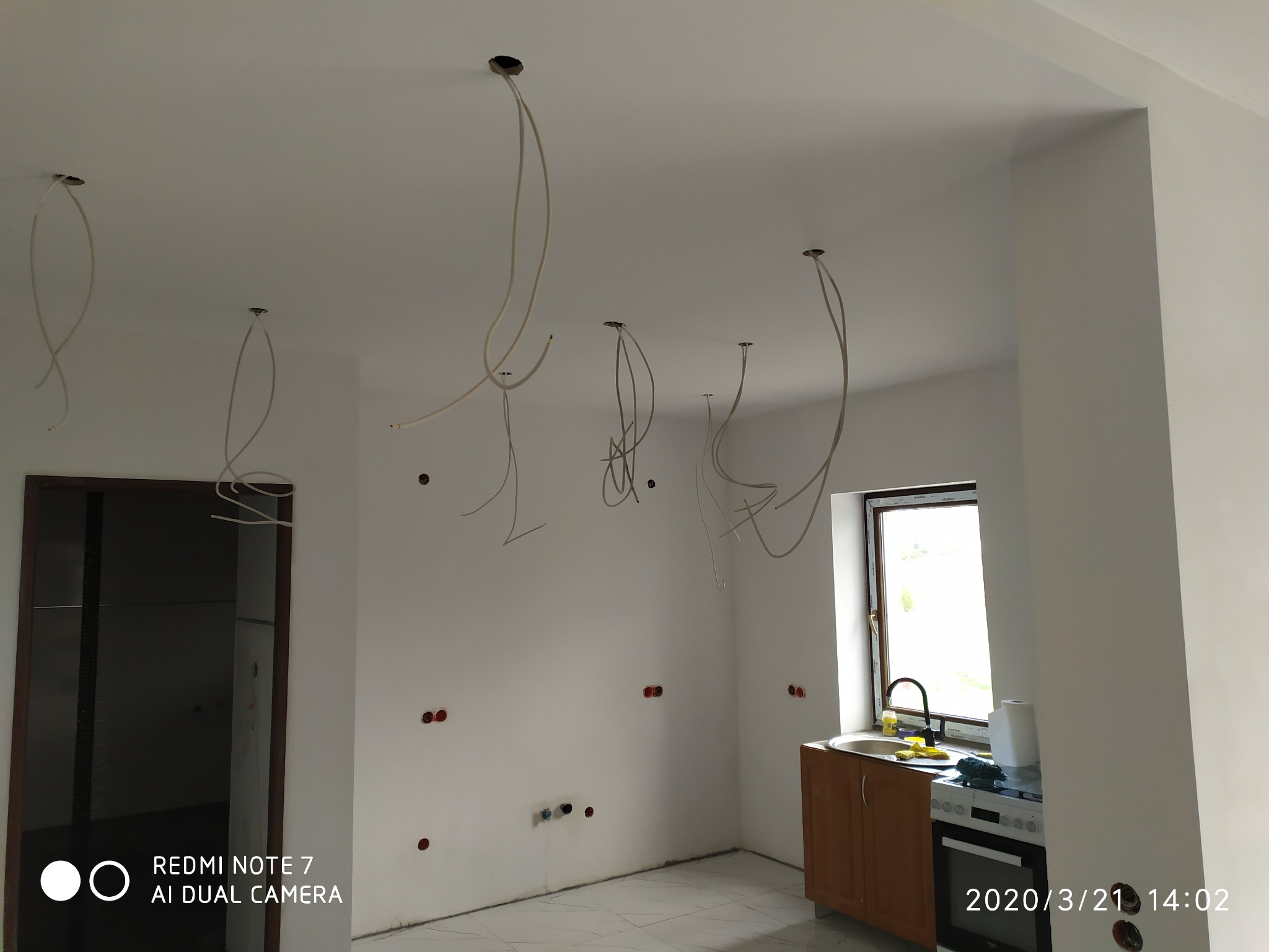 Wnętrze w trakcie prac instalacyjnych: widoczne wystające kable z sufitu, puszki elektryczne w ścianie, fragment kuchni z oknem w tle.