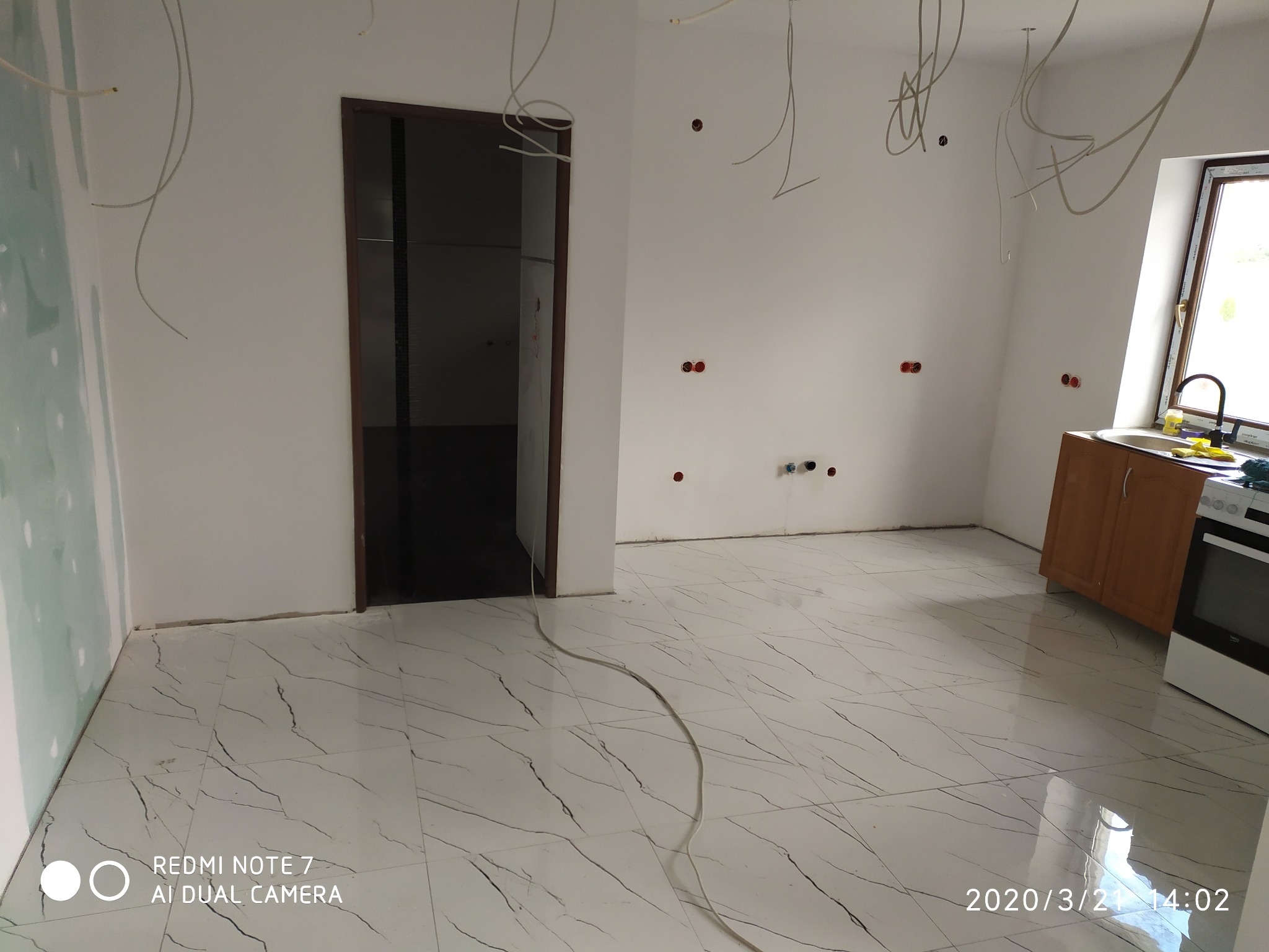 Wnętrze w trakcie prac wykończeniowych: białe ściany z wystającymi przewodami elektrycznymi, marmurowe płytki podłogowe z czarnymi żyłkami, zamontowane drzwi z ciemną ościeżnicą, fragment szafki...