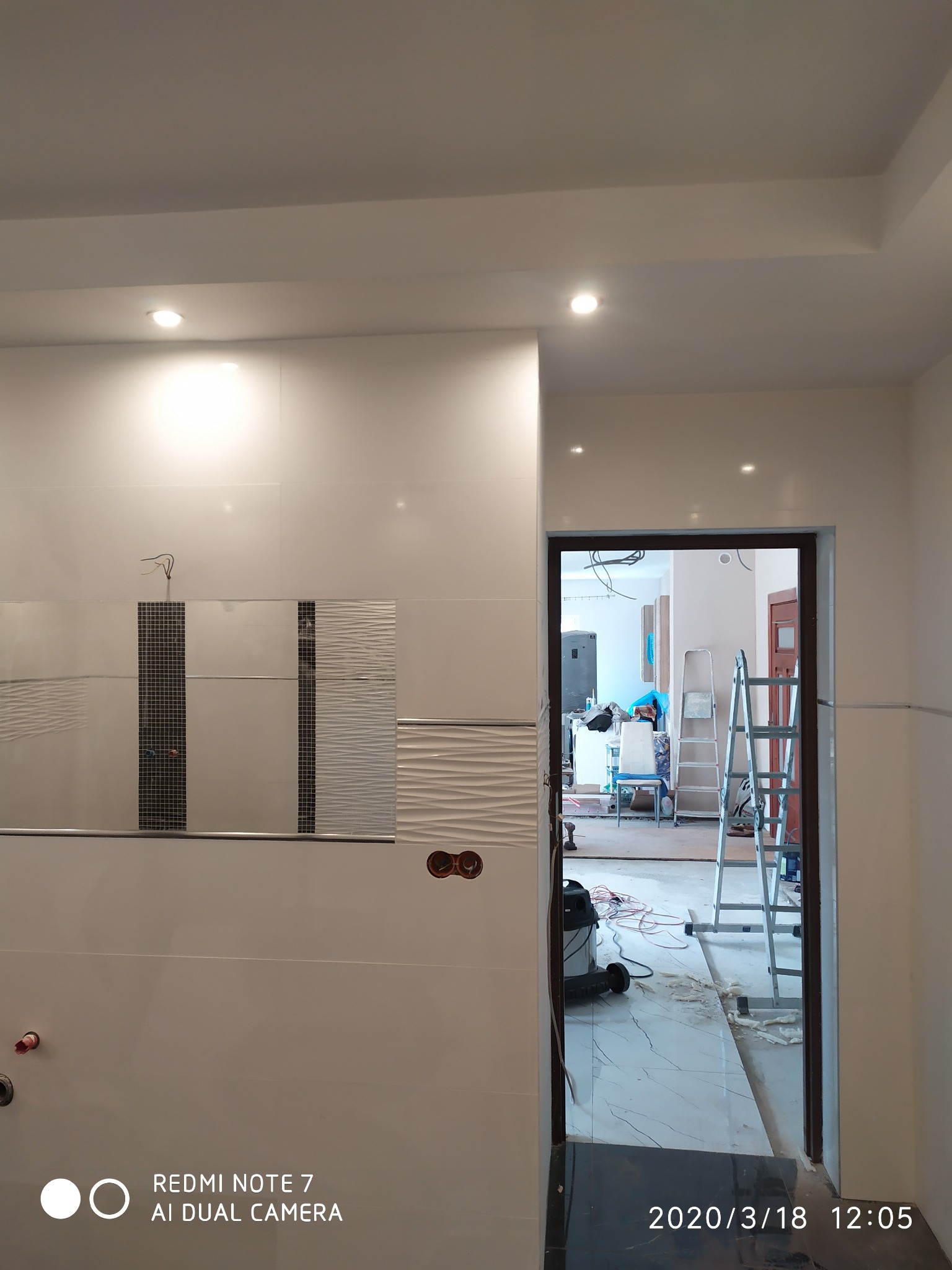 Wnętrze łazienki w trakcie prac wykończeniowych: białe płytki z dekoracyjnymi wstawkami, wystające kable, widoczne otwory na gniazdka, przez otwarte drzwi widoczny bałagan i drabina.