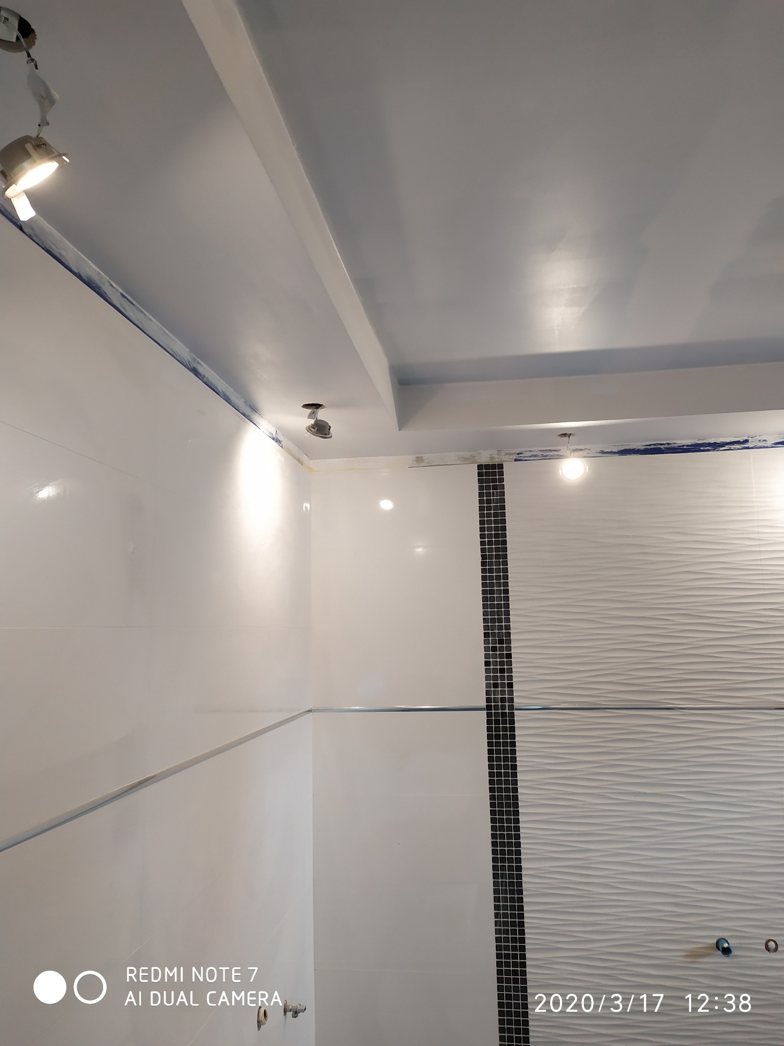 Białe, błyszczące płytki na ścianach wykończonej łazienki, z dekoracyjną wstawką z czarno-białej mozaiki i płytkami z falistą strukturą, oświetlone punktowymi lampami sufitowymi.