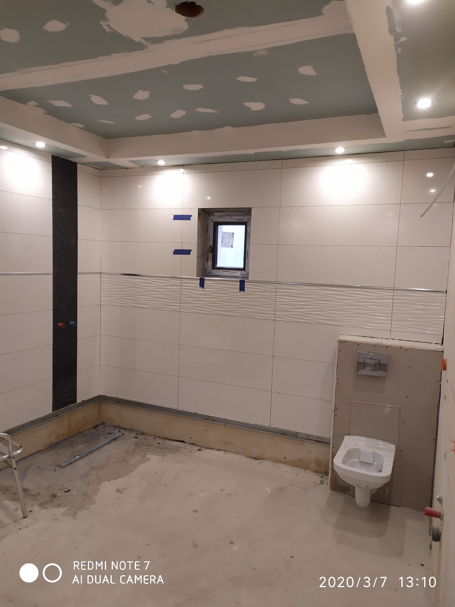 Łazienka w trakcie remontu: białe płytki z poziomym wzorem, czarny pas mozaiki, częściowo wykończony sufit z oświetleniem punktowym, zamontowana toaleta, okno zabezpieczone taśmą malarską.