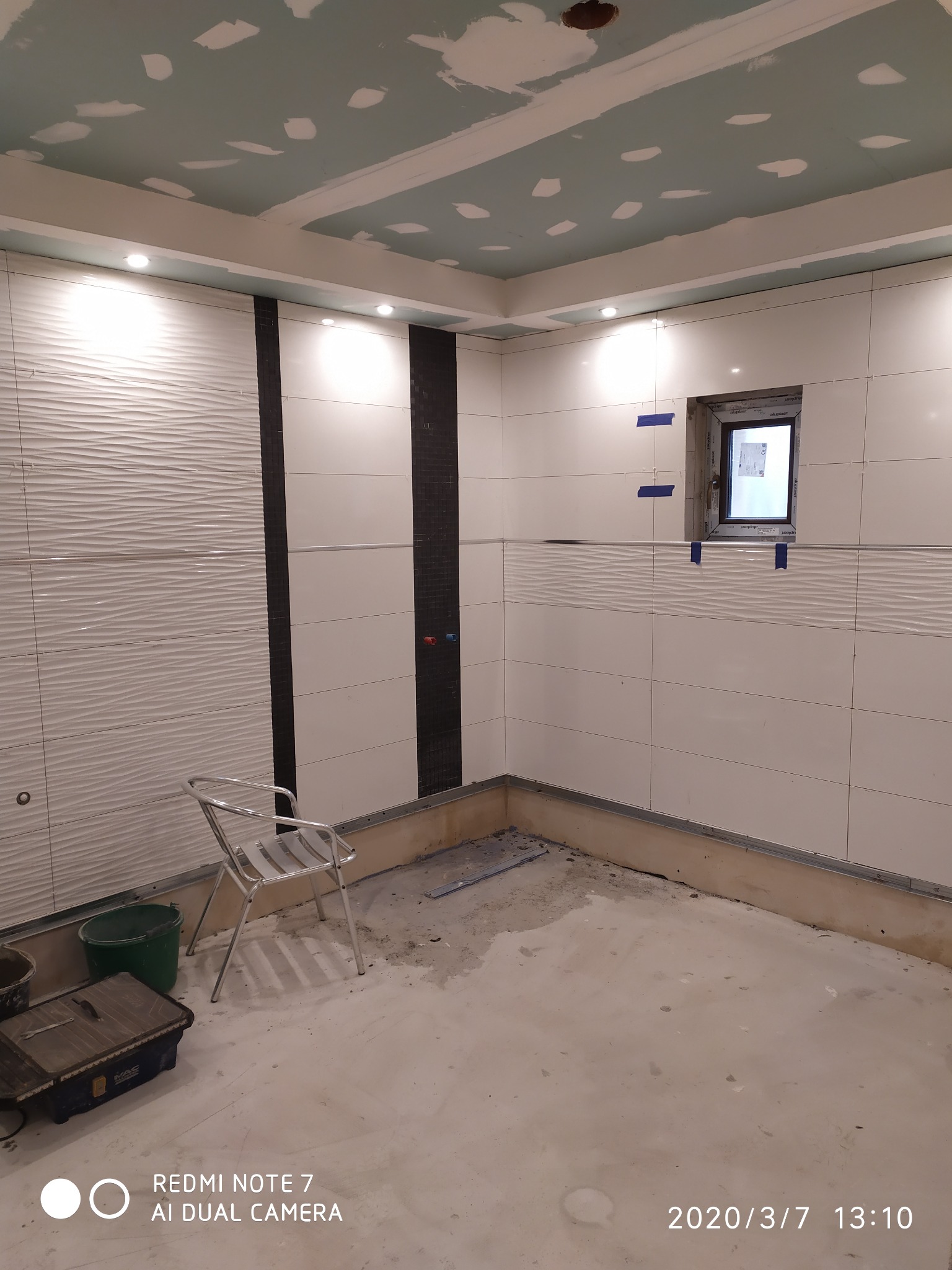 Wnętrze łazienki w trakcie remontu: białe płytki z poziomym wzorem fal, czarny pas płytek, okno zabezpieczone taśmą, sufit z płyt gipsowo-kartonowych przed szpachlowaniem, metalowe krzesło...