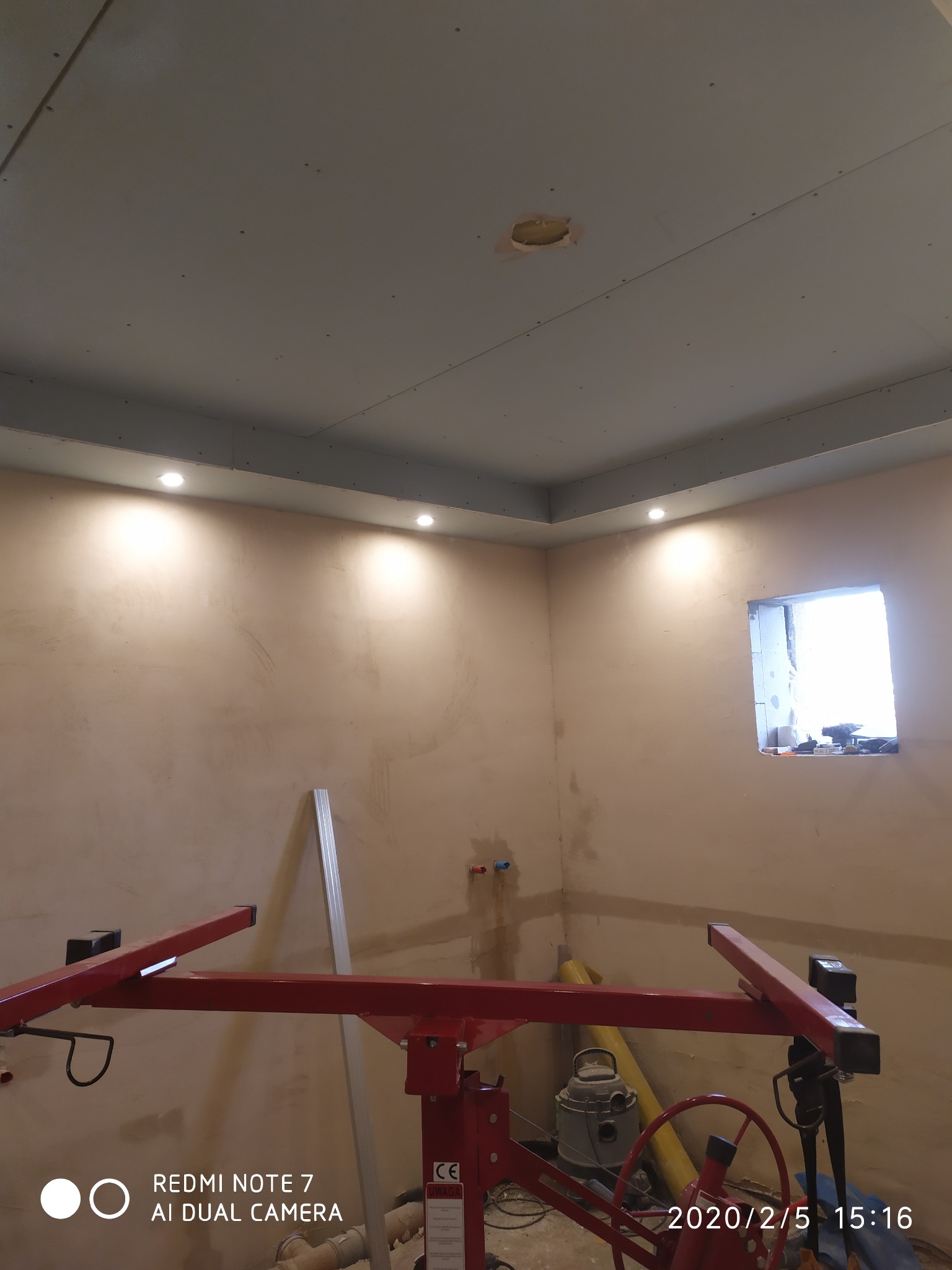 Wnętrze w trakcie prac wykończeniowych: sufit z płyt gipsowo-kartonowych z wbudowanymi punktami świetlnymi, ściany w stanie surowym, widoczny podnośnik do płyt i okno w trakcie montażu.