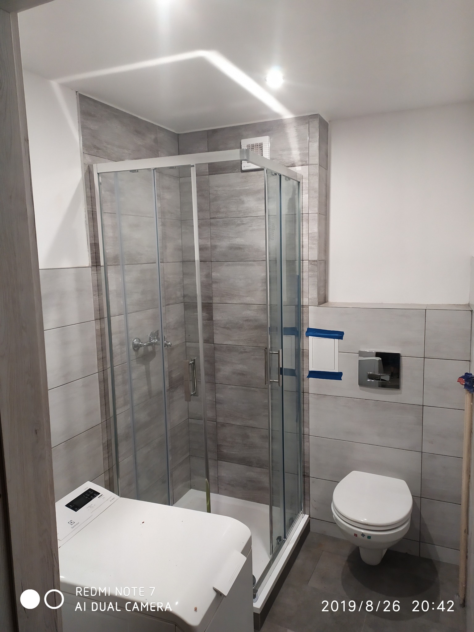 Wykończona łazienka z kabiną prysznicową, toaletą i pralką, wyłożona szarymi płytkami imitującymi kamień, z widocznymi elementami instalacji zabezpieczonymi taśmą malarską.