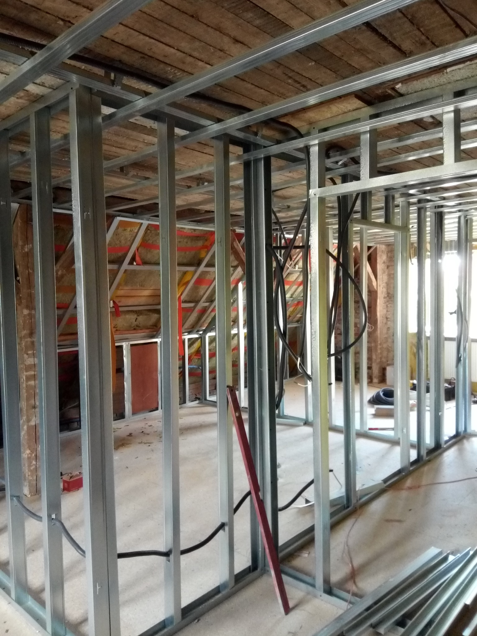 Konstrukcja ścian działowych z profili stalowych w surowym stanie, widoczne przewody elektryczne, drewniany strop w tle, etap prac wykończeniowych poddasza.