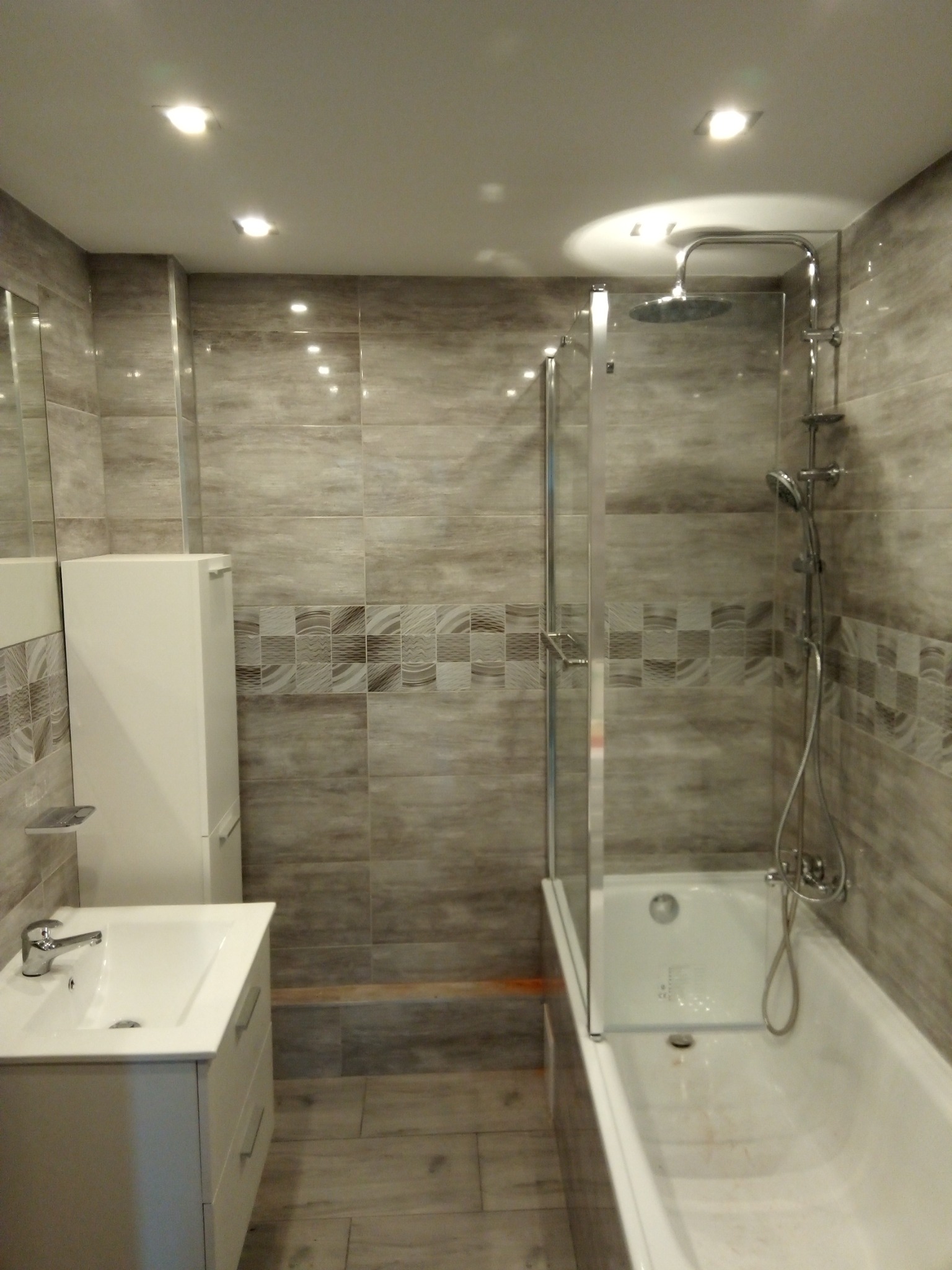 Wykończona łazienka z wanną z parawanem, umywalką z szafką i płytkami w odcieniach szarości z dekoracyjnym pasem mozaiki.