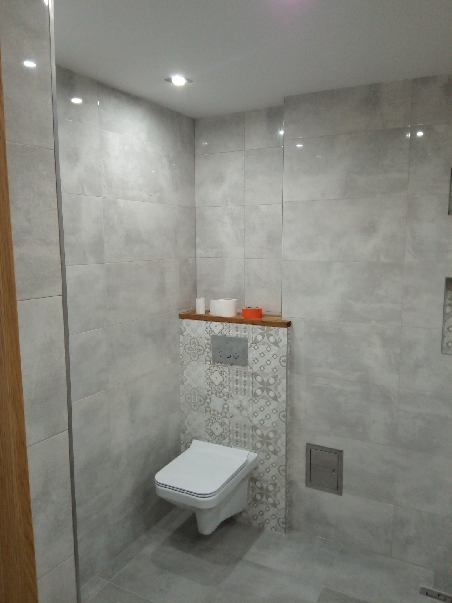 Nowoczesna łazienka z szarymi płytkami imitującymi kamień, wiszącą toaletą i dekoracyjną ścianką nad nią, drewniana półka z akcesoriami łazienkowymi.