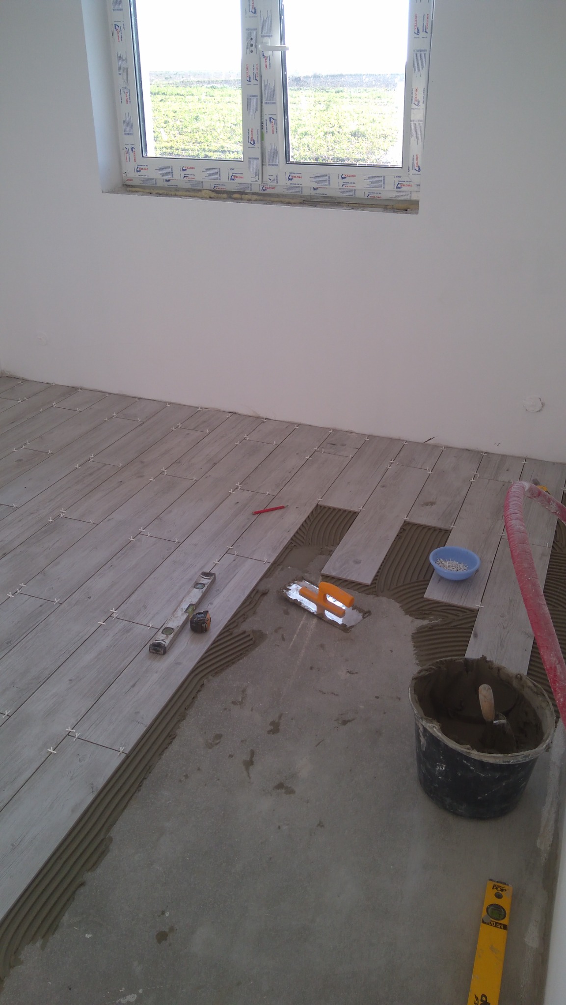Układanie podłogi z płytek imitujących drewno w jasnym odcieniu, widoczne narzędzia: poziomica, kielnia, krzyżyki dystansowe, wiadro z zaprawą, czerwony przewód i okno w tle.