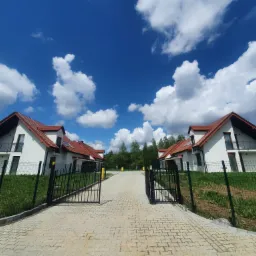 Nowo wybudowane, białe domy jednorodzinne z czerwoną dachówką, widziane z perspektywy wjazdu przez otwartą, czarną bramę, na tle błękitnego nieba z białymi chmurami.