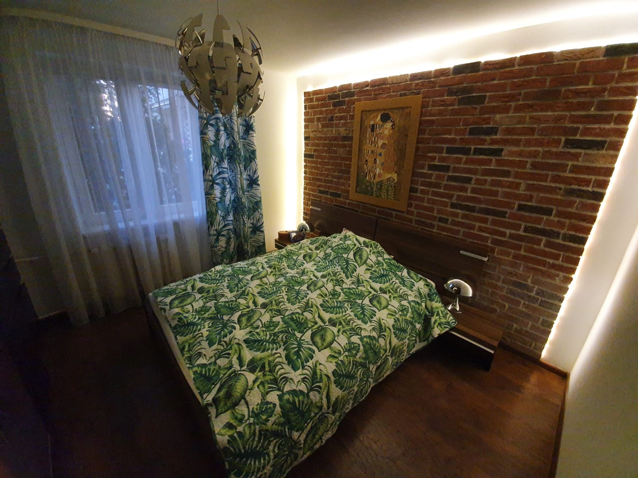 Sypialnia z łóżkiem w zielone liście, ceglaną ścianą z podświetleniem LED i obrazem w złotej ramie, lampa wisząca w stylu nowoczesnym i zasłony w tropikalny wzór.