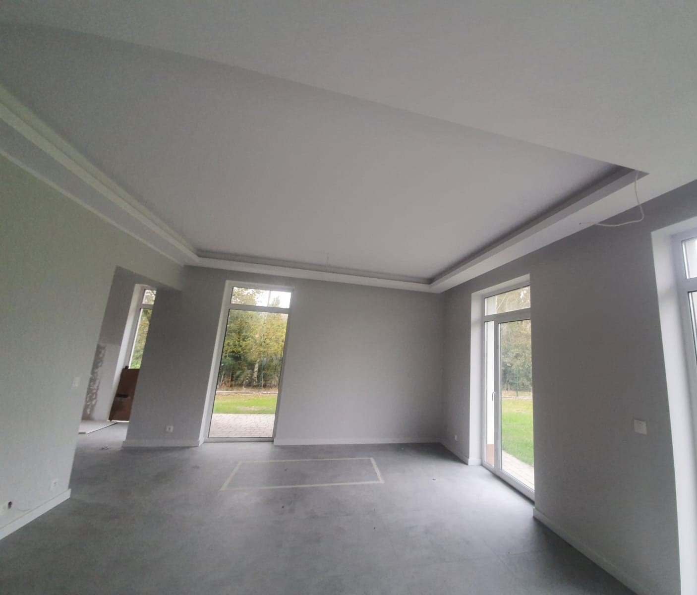 Szare wnętrze w trakcie wykończenia z białym sufitem podwieszanym z dekoracyjnymi listwami i dużymi oknami z widokiem na zieleń.