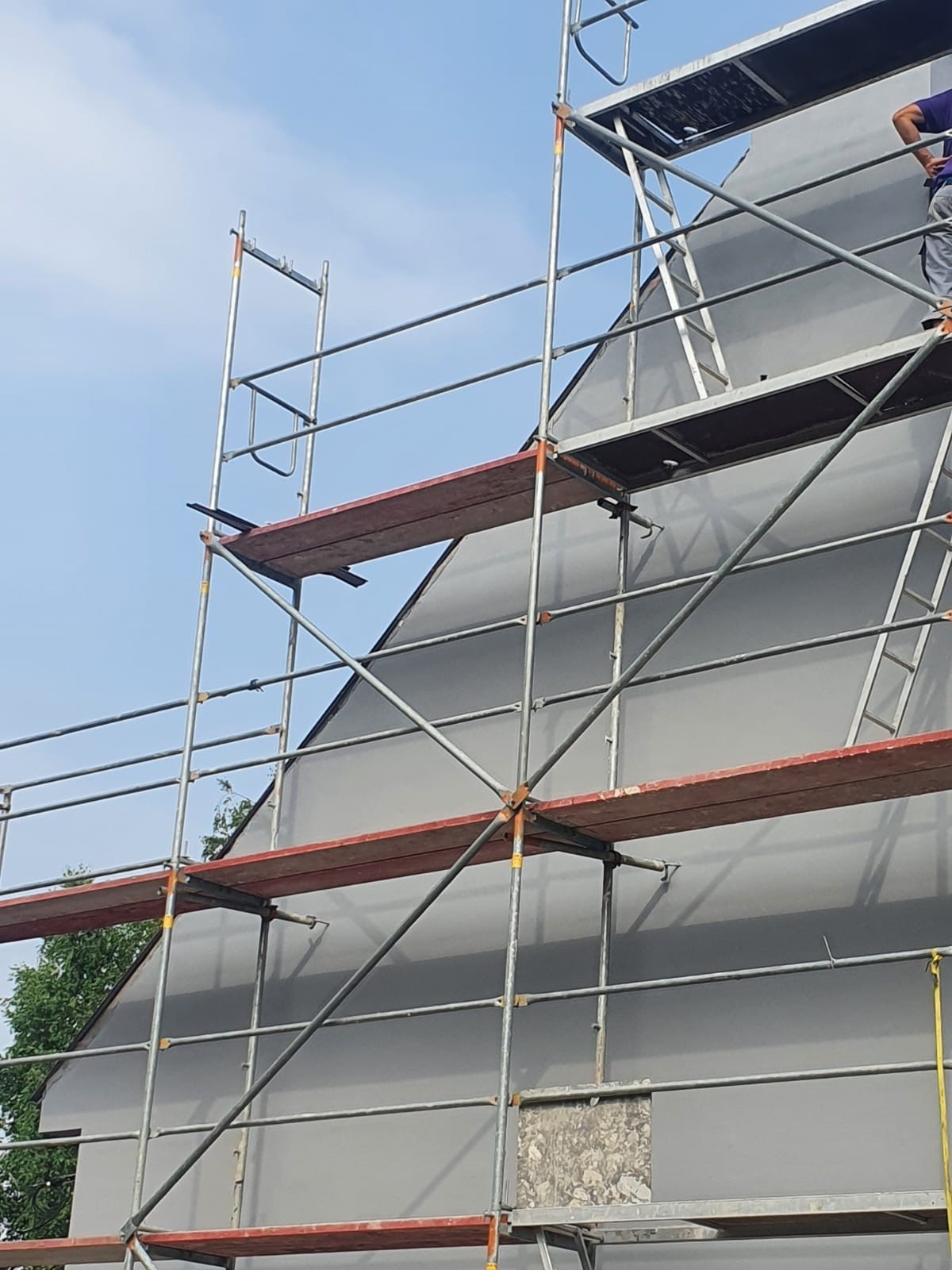 Dom w trakcie ocieplania, widoczny fragment elewacji pokryty szarym materiałem izolacyjnym i rusztowanie, w tle błękitne niebo.