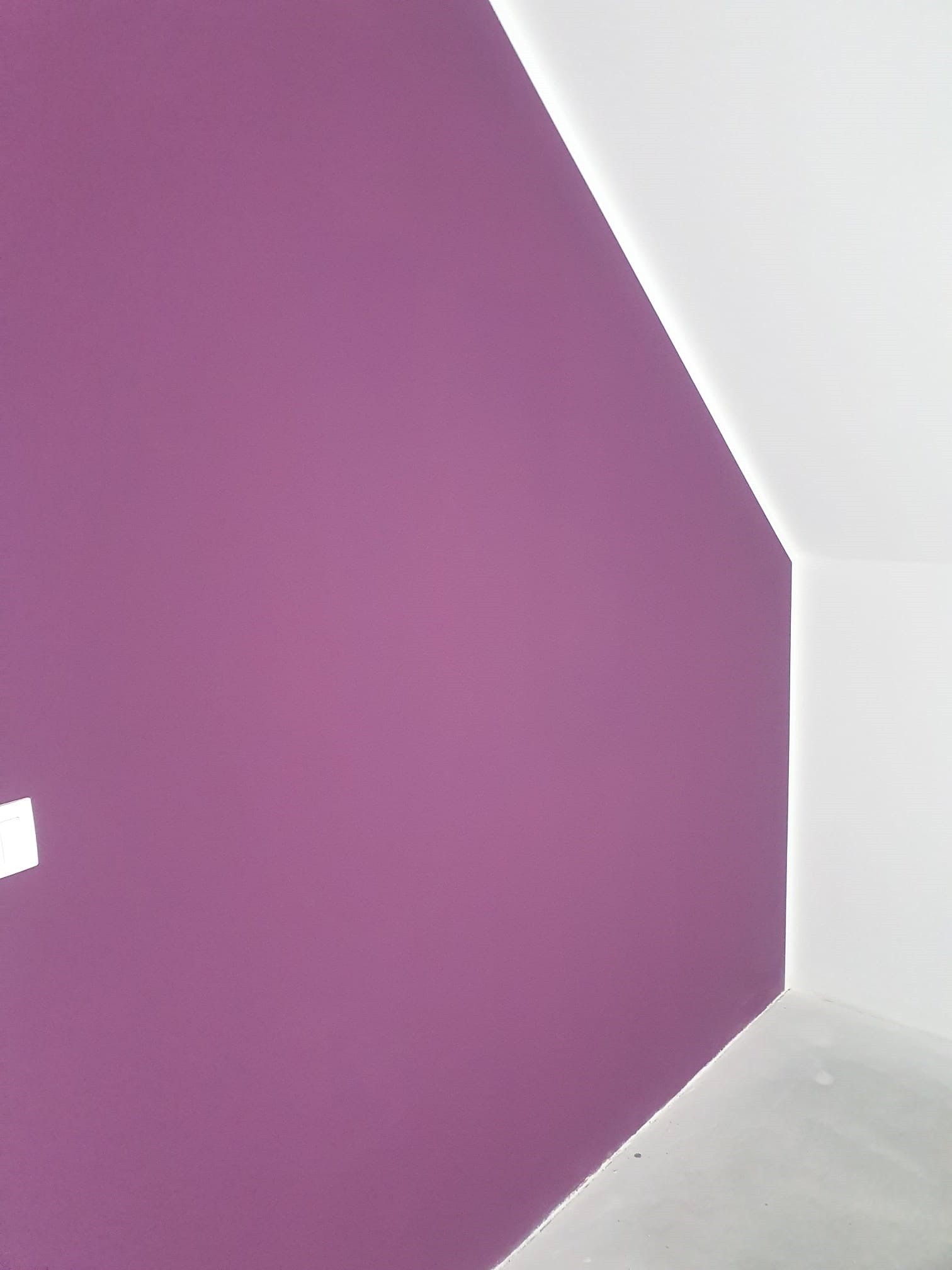 Świeżo pomalowana ściana w odcieniu fioletu w rogu pokoju, kontrastująca z białym sufitem i fragmentem podłogi.