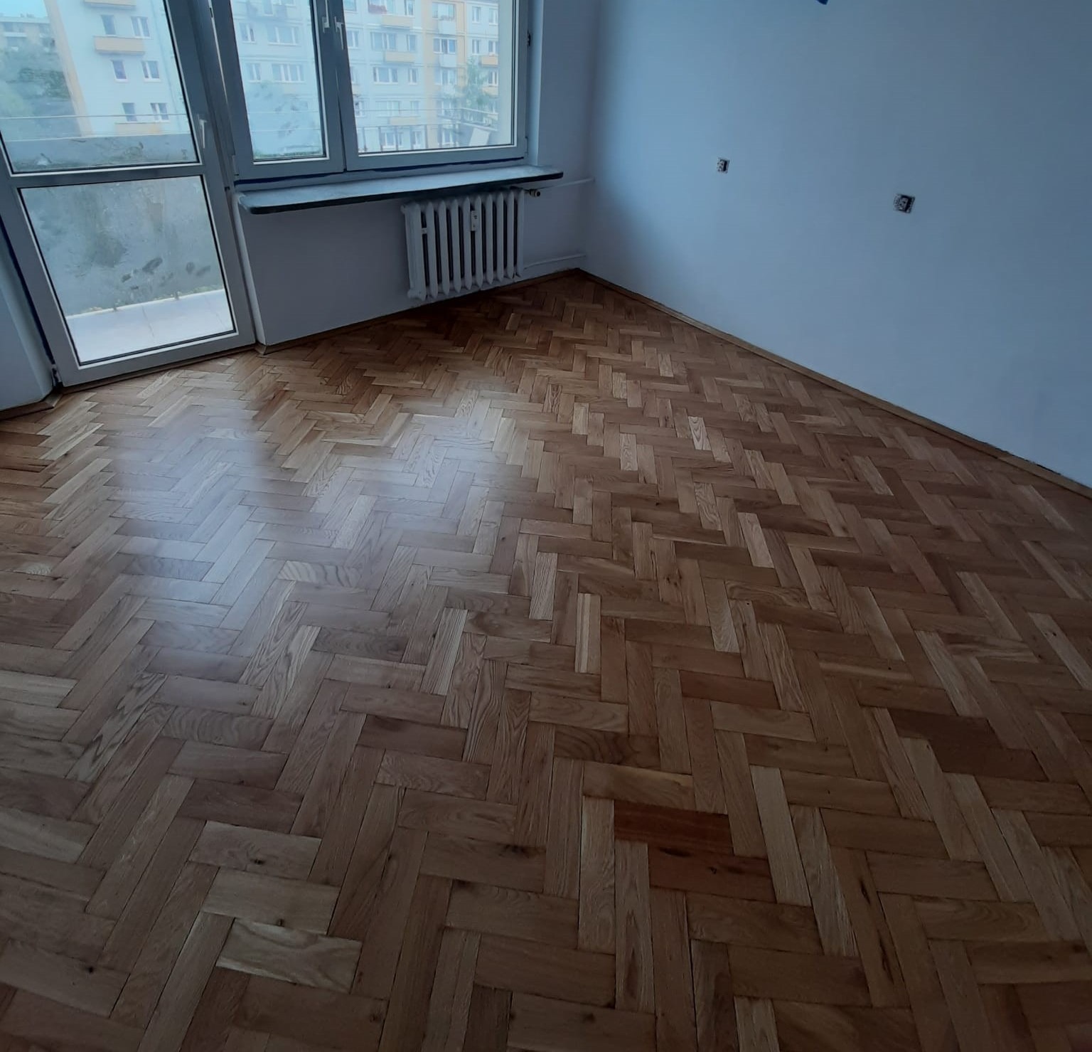 Ułożony parkiet w jodełkę w pustym pokoju z oknem balkonowym i grzejnikiem, widoczne gniazdka elektryczne na ścianie.