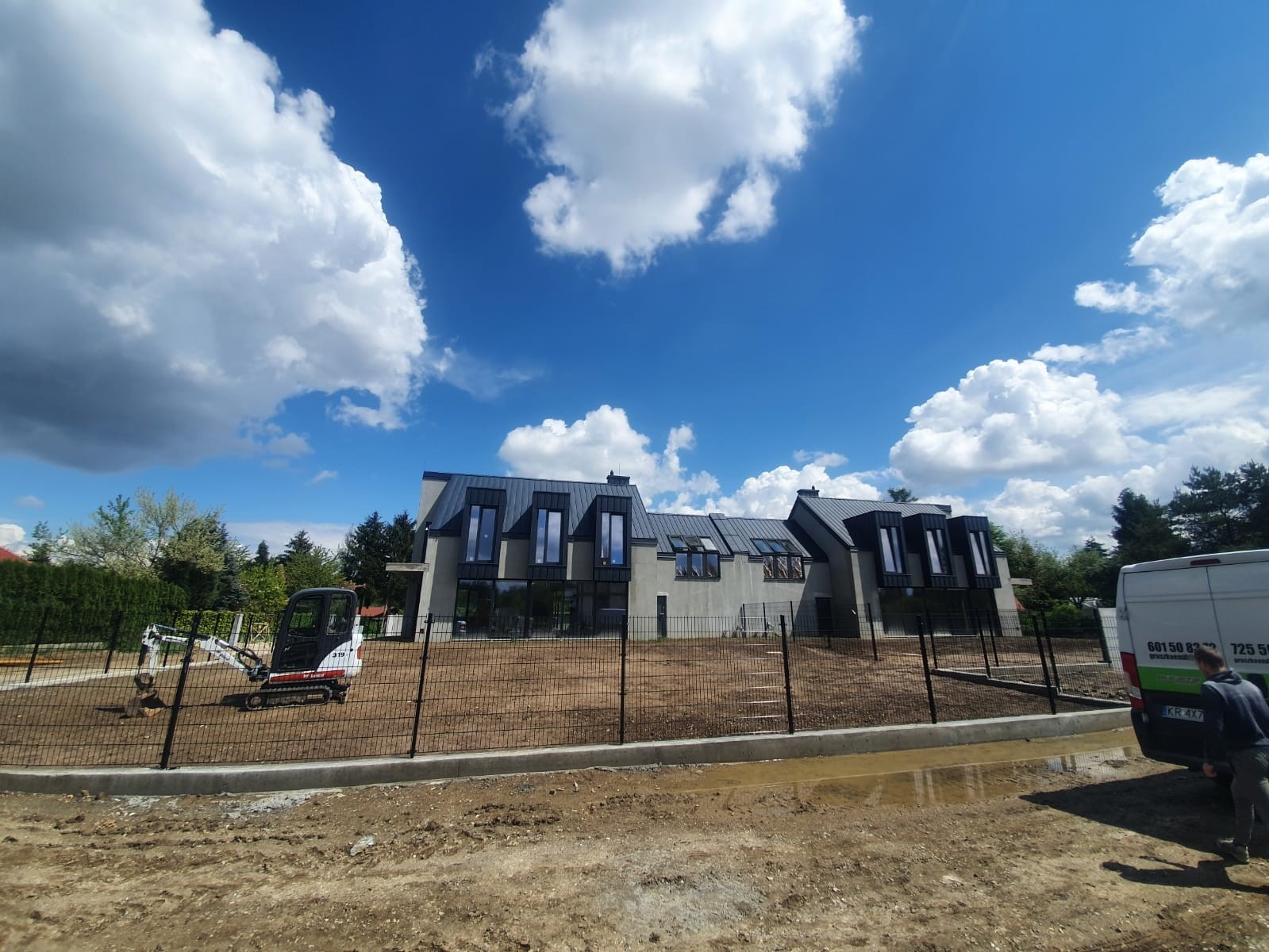 Nowoczesny dom jednorodzinny z szarymi elewacjami i czarnymi oknami, ogrodzony metalową siatką, z małą koparką na terenie działki, widok na błękitne niebo z obłokami.