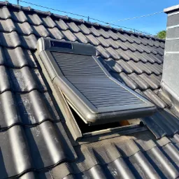 Otwarta roleta zewnętrzna na oknie dachowym, zamontowana na ciemnym, ceramicznym dachu w słoneczny dzień.
