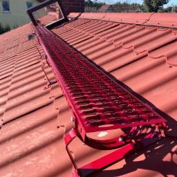 Czerwona ława kominiarska zamontowana na czerwonej dachówce, z widocznym oknem dachowym i kominem w tle, oświetlone jasnym słońcem.