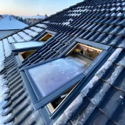 Dwa otwarte okna dachowe Velux na ciemnym dachu pokrytym dach&oacute;wką, częściowo ośnieżonym, widok z dołu pod kątem, w tle zabudowa jednorodzinna.