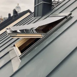 Szare okno dachowe z częściowo rozłożoną, zaciemniającą roletą na metalowym dachu, widok z góry, na dachu widoczne anteny satelitarne.