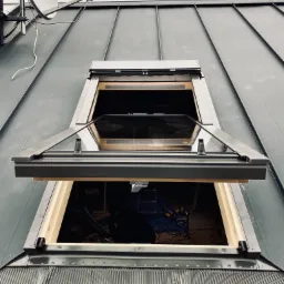 Otwarty okno dachowe z widokiem na wnętrze poddasza podczas montażu, widoczna antena satelitarna na dachu z blachy trapezowej.