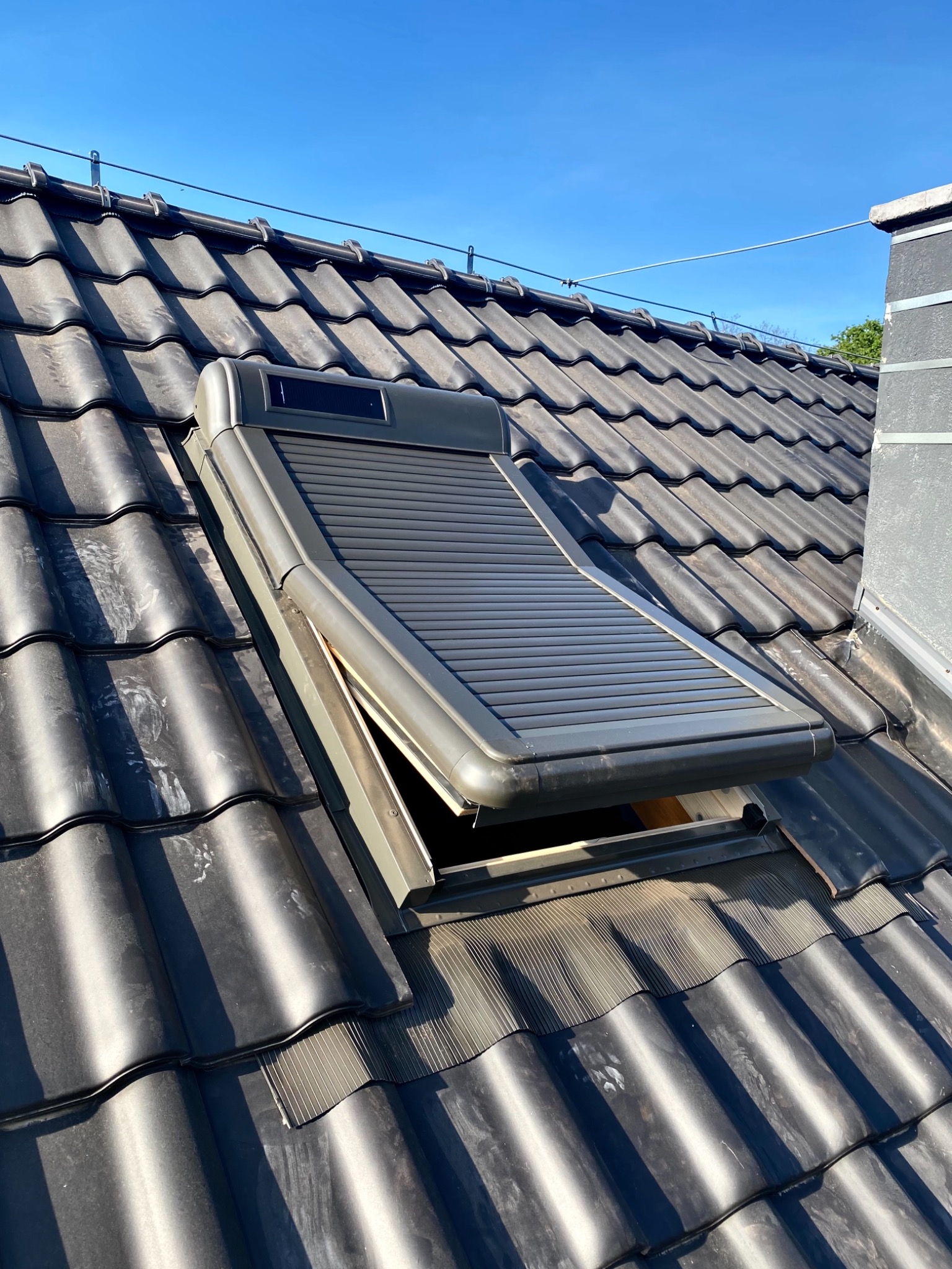 Otwarta roleta zewnętrzna na oknie dachowym, zamontowana na ciemnym, ceramicznym dachu w słoneczny dzień.