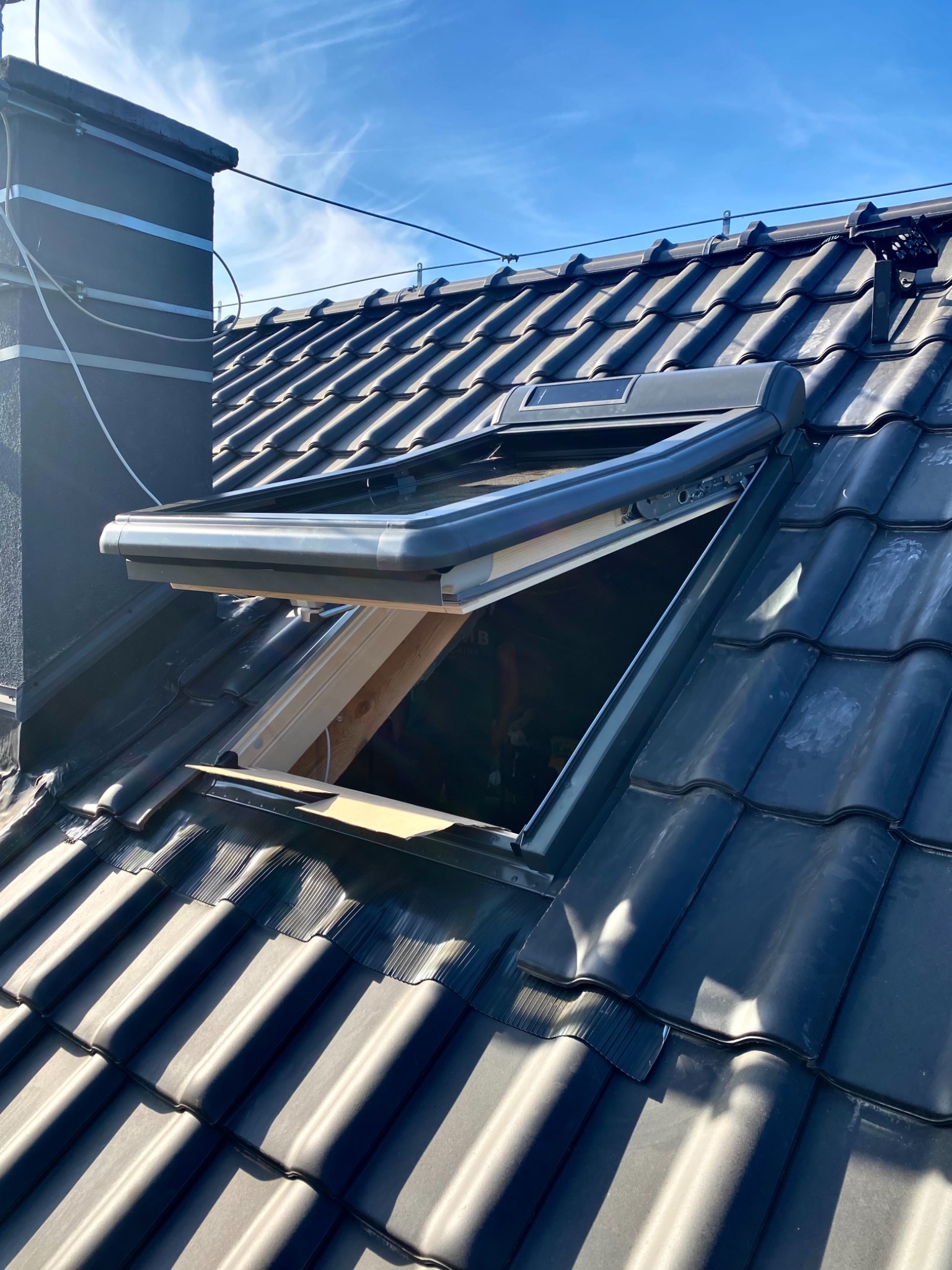 Otwarty, czarny okno dachowe na pokrytym ciemną dachówką dachu, widok z dołu na błękitne niebo, fragment komina w tle.