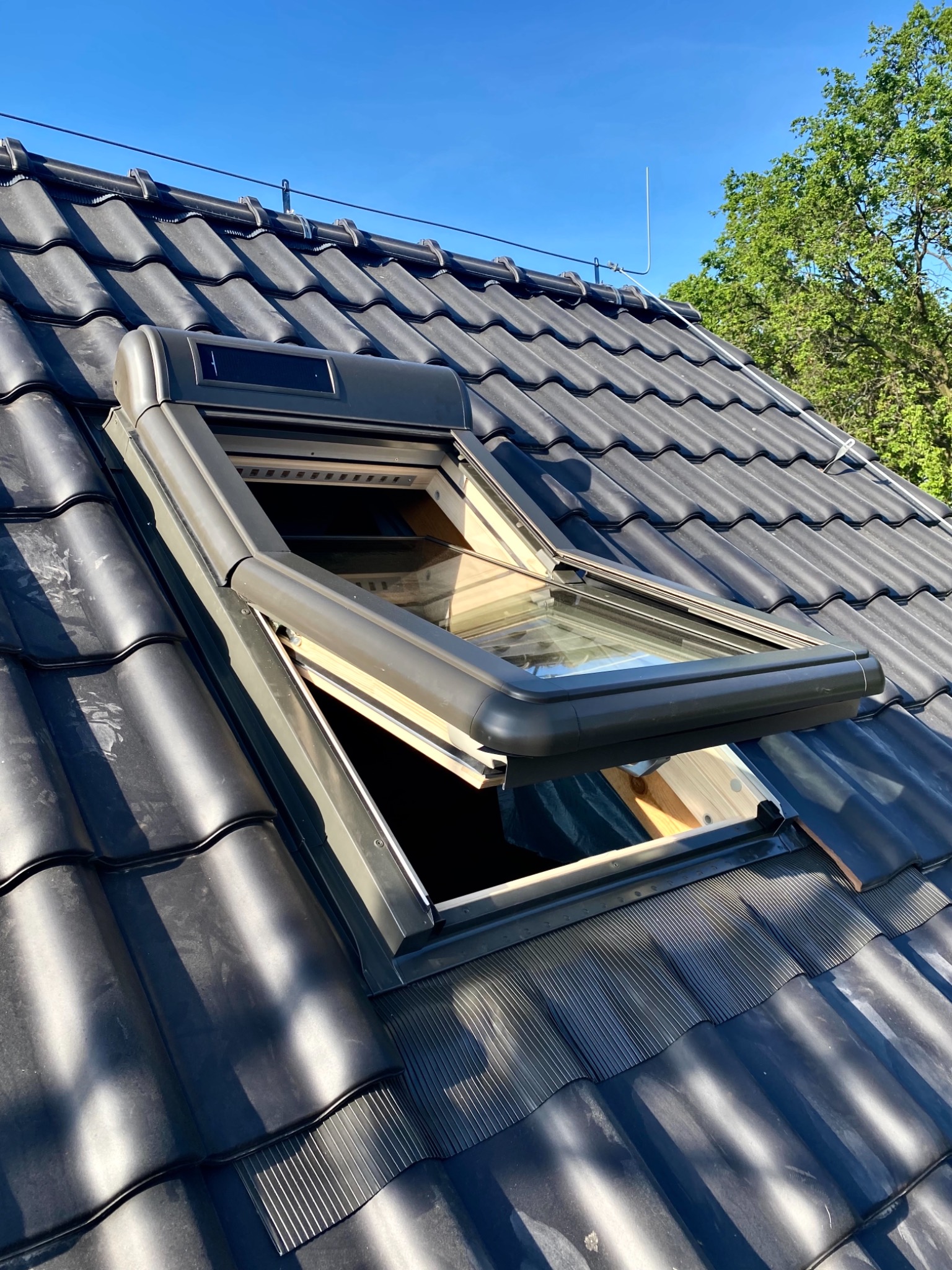 Otwarty, uchylny lukarnowy system okienny wkomponowany w ciemną dachówkę, widoczna konstrukcja ramy okna i jasne niebo w tle.