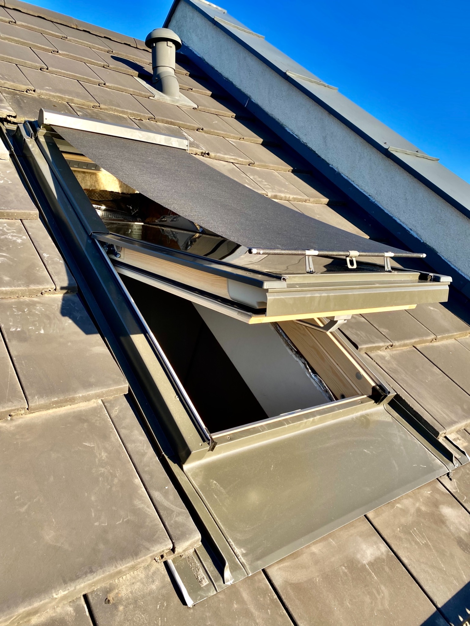 Otwarty lukarn z zamontowaną, częściowo zwiniętą roletą zaciemniającą, widok z góry na dach pokryty dachówką ceramiczną w słoneczny dzień.