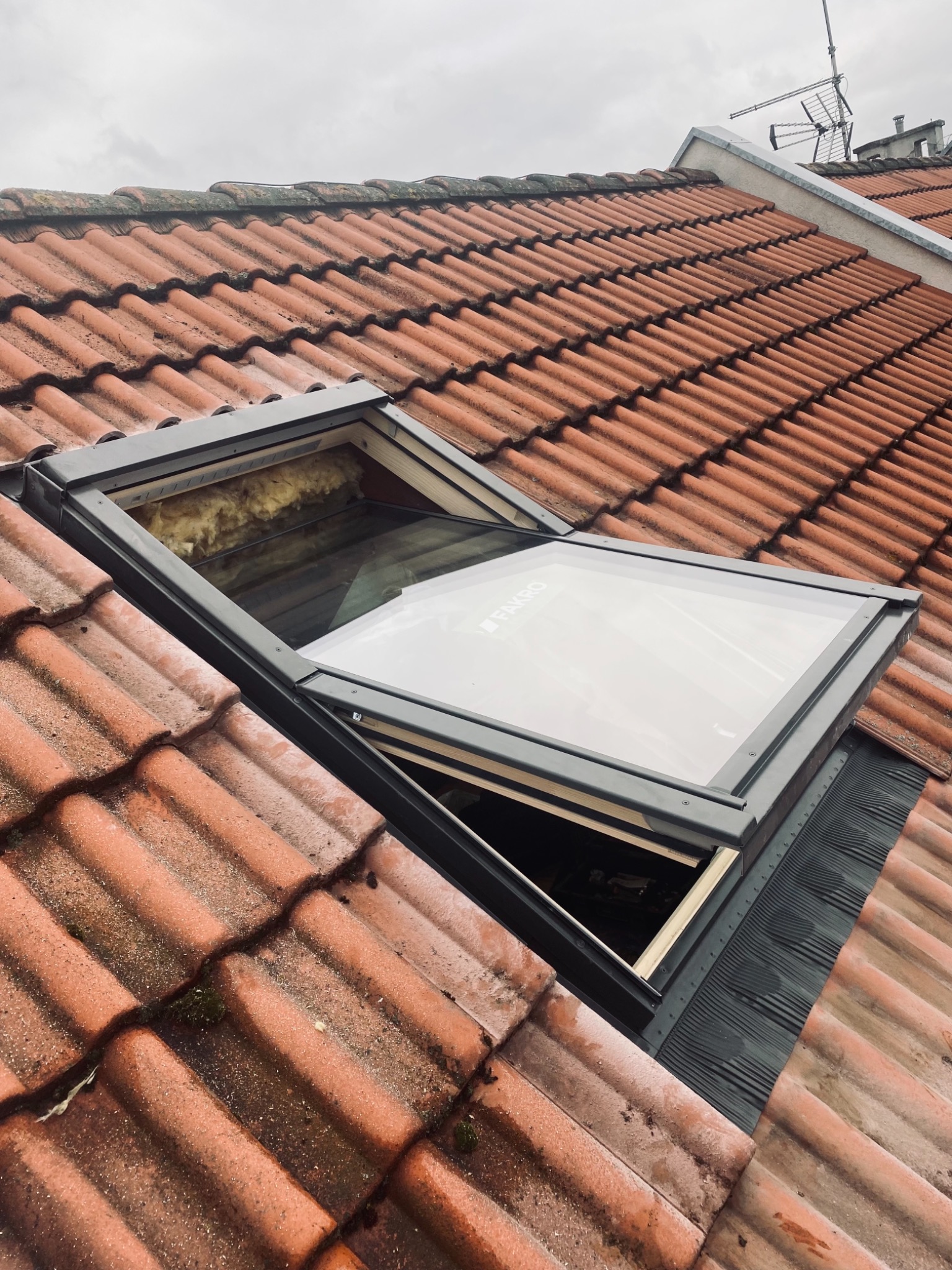 Otwarty, szary dachowy element wbudowany w czerwoną dachówkę, widoczne ocieplenie i logo Fakro na szybie.