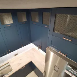 Niebieska zabudowa kuchenna z drewnianymi blatami, siatkowymi frontami szafek górnych i miedzianymi uchwytami, widok z góry.