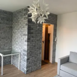 Wykończone wnętrze mieszkania z szarą cegłą dekoracyjną na ścianie, nowoczesną lampą sufitową w kształcie kwiatu i stolikiem ze szklanym blatem, widoczne fragment sofy i drabina malarska w tle.