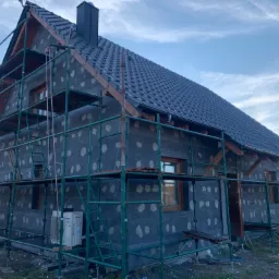 Dom w trakcie ocieplania, widoczne rusztowania i warstwa izolacyjna z plackami kleju, dach pokryty ciemną dachówką, błękitne niebo w tle.