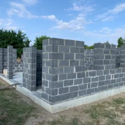 Wznoszone ściany parteru domu z bloczk&oacute;w betonowych na fundamencie, widok z zewnątrz w słoneczny dzień.