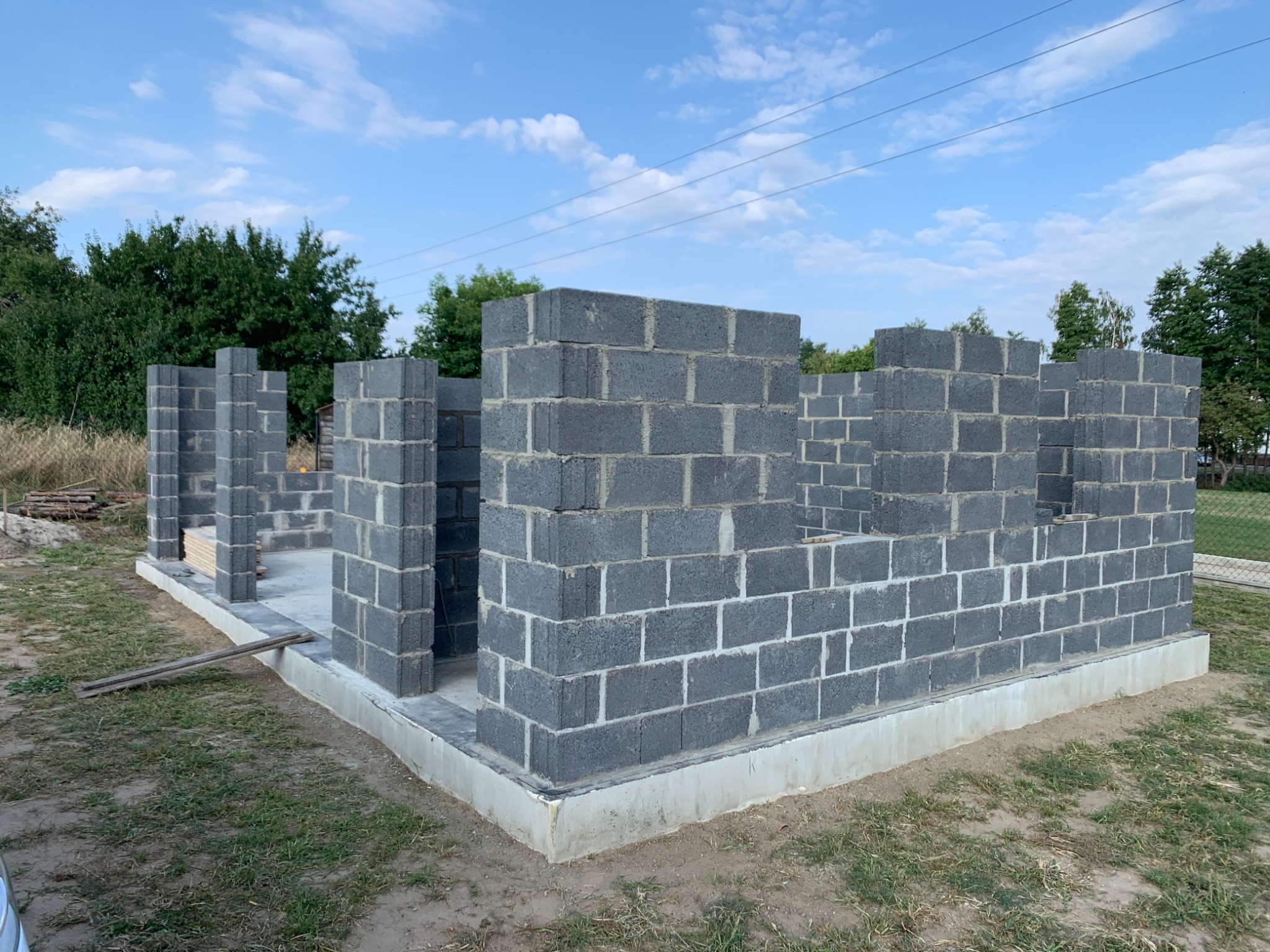 Wznoszone ściany parteru domu z bloczków betonowych na fundamencie, widok z zewnątrz w słoneczny dzień.