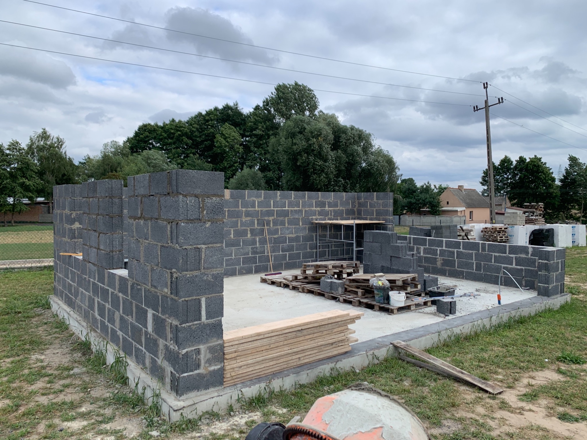 Wznoszenie ścian parteru domu z bloczków betonowych na wylanym fundamencie, widoczne palety z bloczkami i deski, w tle drzewa i pochmurne niebo.
