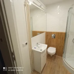 Wykończona łazienka z umywalką na szafce, toaletą i kabiną prysznicową, białe i drewnopodobne płytki na ścianach, widoczne rury grzewcze.