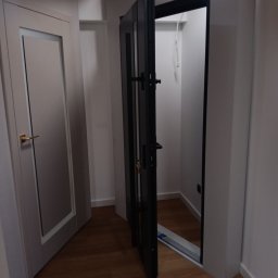 J.FELIŃSKI OKNA DRZWI ROLETY USŁUGI REMONTOWE JANUSZ FELIŃSKI - Ciemne, nowoczesne drzwi otwarte do garderoby, widoczne na tle drewnianej podłogi i białych ścian. Obok drzwi z przeszkleniem i złotą klamką. Widoczny fragment drewnianej belki stropowej.