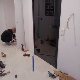 J.FELIŃSKI OKNA DRZWI ROLETY USŁUGI REMONTOWE JANUSZ FELIŃSKI - Montaż czarnych, szklanych drzwi wewnętrznych. Widoczne narzędzia, w tle osoba przy białej ścianie. Na podłodze resztki materiałów i akcesoriów montażowych.