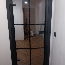 J.FELIŃSKI OKNA DRZWI ROLETY USŁUGI REMONTOWE JANUSZ FELIŃSKI - Nowoczesne drzwi wewnętrzne z czarną ramą i szklanym wypełnieniem, widok na parkiet ułożony w jodełkę. Minimalistyczny design, jasne ściany.