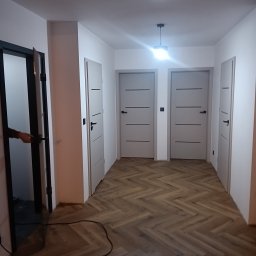 J.FELIŃSKI OKNA DRZWI ROLETY USŁUGI REMONTOWE JANUSZ FELIŃSKI - Jasne, minimalistyczne wnętrze z trzema parami drzwi w odcieniach szarości i podłogą ułożoną w jodełkę. Jedne drzwi są otwarte, ukazując fragment dłoni na klamce.